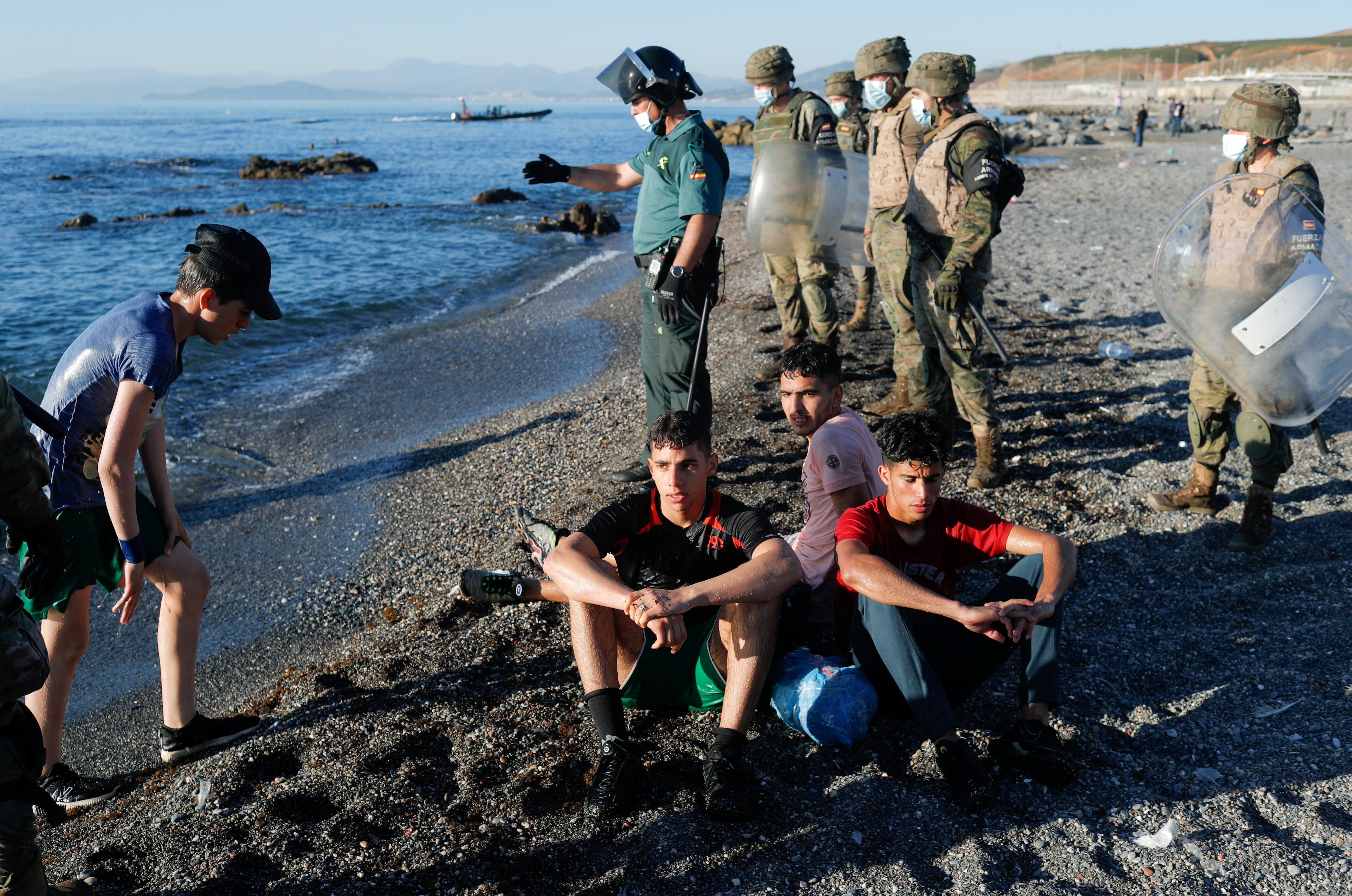 Spanish soldiers stand guard as Moroccan citizens sit at El Tarajal beach, near the fence between the Spanish-Moroccan border, after thousands of migrants swam across the border, in Ceuta, Spain, May 19, 2021. REUTERS/Jon Nazca
