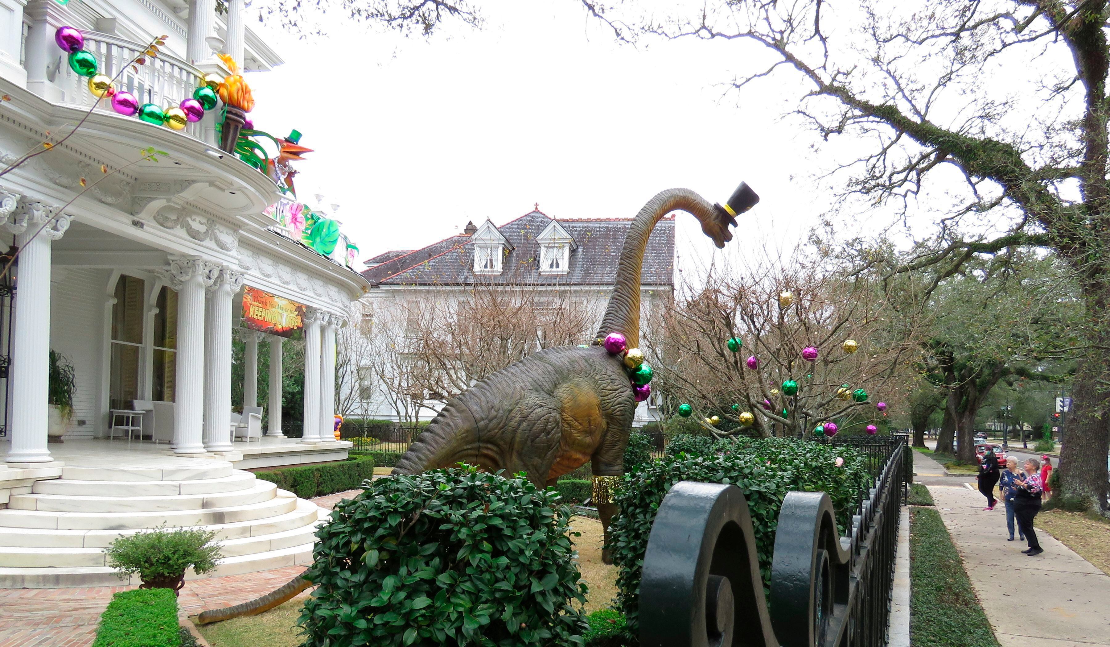 Transeúntes se detienen a ver un enorme dinosaurio en una mansión en la avenida St. Charles de Nueva Orleans el martes 26 de enero de 2021. La pancarta dice: "Gracias, alcaldesa, por mantenernos seguros". Con la cancelación de los tradicionales desfiles de Mardi Gras debido a la pandemia de coronavirus, miles alrededor de la ciudad han decorado sus casas para celebrar el Carnaval. (AP Foto/Janet McConnaughey)