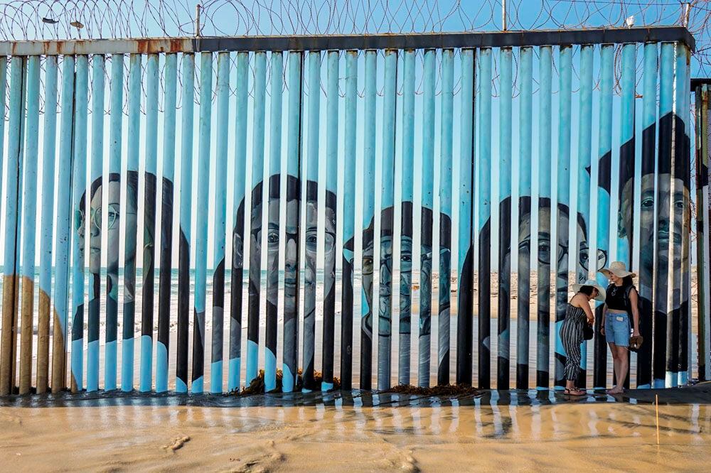 Personas caminan hoy junto a un mural con los rostros de personas deportadas pintado sobre la barrera metálica fronteriza en Tijuana, estado de Baja California (México). EFE/Joebeth Terriquez
