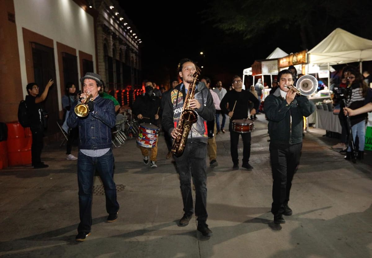Integrantes de una banda musical caminan tocando instrumentos de viento y percusión durante un evento nocturno al aire libre. | Cortesía