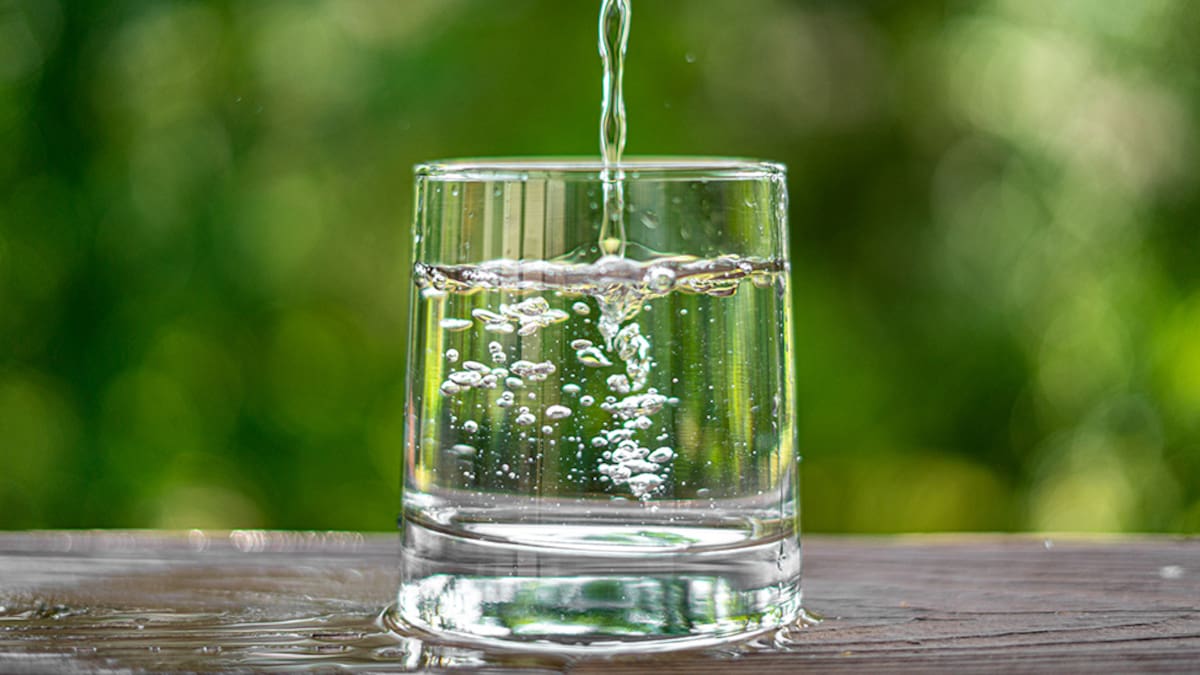 Perfectly vertical stream of water frozen in time hitting a bubbling glass of water sitting outside on the wooden railing of a patio with beautiful bokeh of trees and foliage in the background.