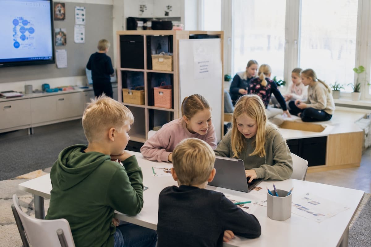 Niños en un aula finlandesa colaborando en actividades prácticas y juegos educativos, con mesas bajas, luz natural y un ambiente moderno que fomenta la creatividad y el aprendizaje activo. | Ilustrativa de ChatGPT