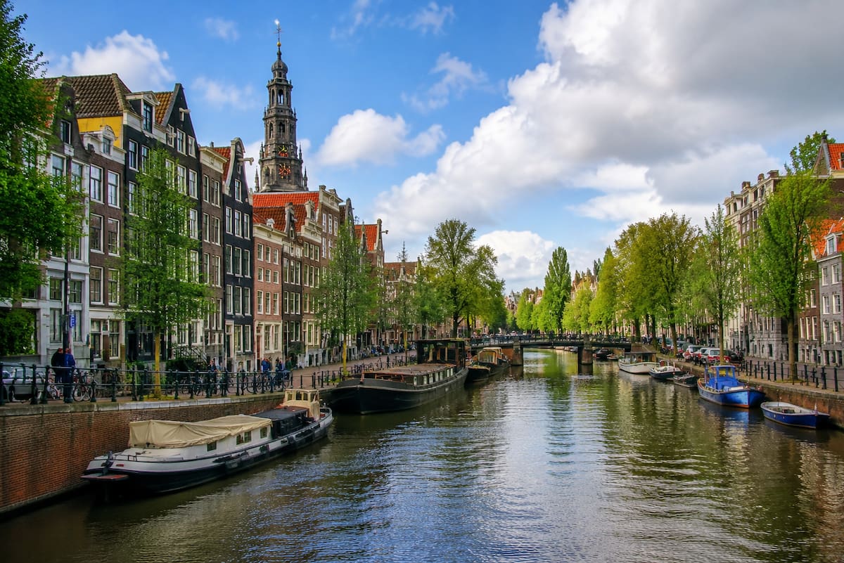 Canal y calles históricas de Ámsterdam, Países Bajos, con casas estrechas de ladrillo, puentes y barcos, mostrando la vida cotidiana junto al agua y la arquitectura típica de la ciudad. | Imagen hecha con IA de ChatGPT