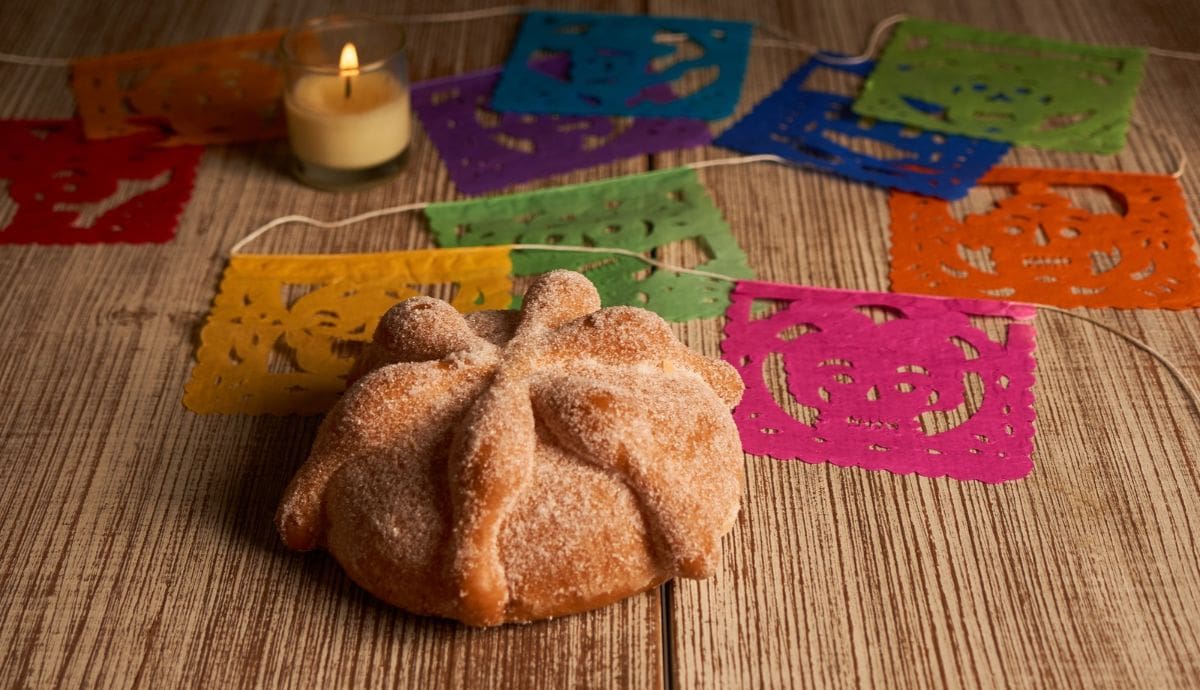 Pan de muerto tradicional espolvoreado con azúcar, acompañado de papel picado multicolor y una vela encendida sobre una superficie de madera durante las festividades de Día de Muertos. | Canva
