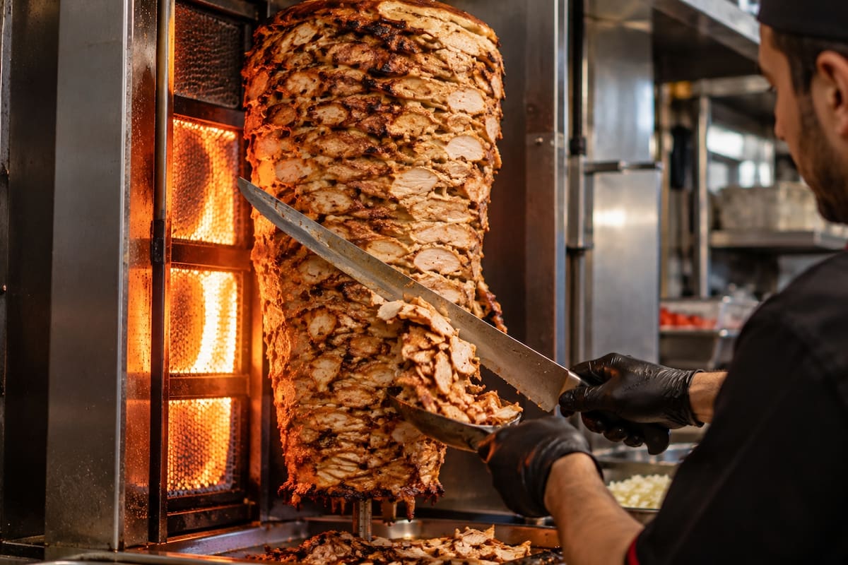 El shawarma se cocina en un asador vertical frente al fuego, donde la carne gira lentamente y se corta en láminas conforme la parte exterior queda dorada. | Crédito: Imagen hecha con IA de ChatGPT