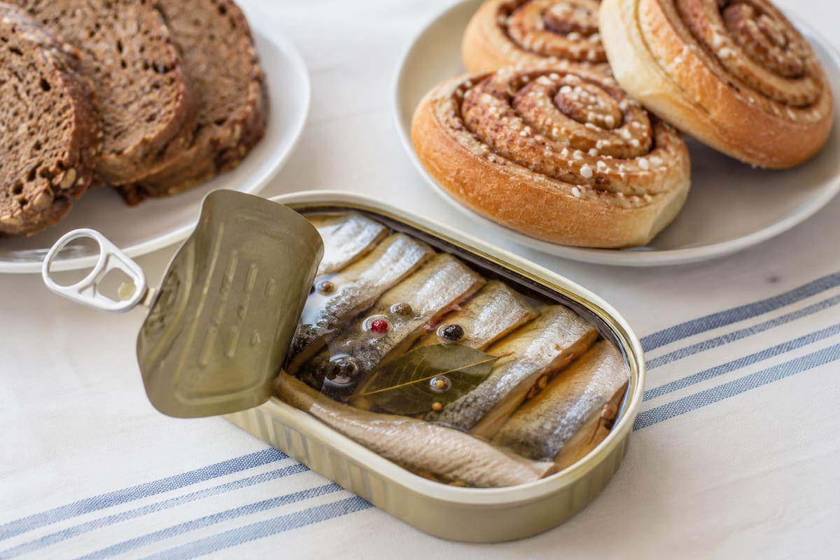 Lata de arenques en conserva acompañada de pan integral y bollos de canela noruegos (kanelboller), mostrando una combinación típica de snacks y bocados tradicionales del norte de Europa. | Imagen hecha con IA de ChatGPT