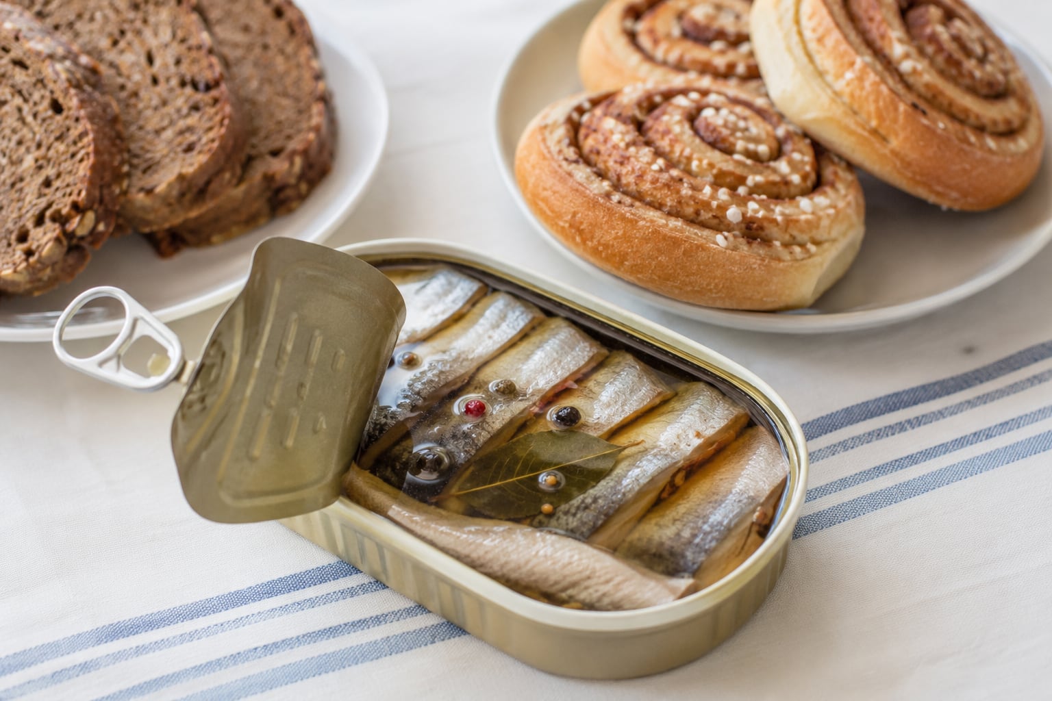 Lata de arenques en conserva acompañada de pan integral y bollos de canela noruegos (kanelboller), mostrando una combinación típica de snacks y bocados tradicionales del norte de Europa. | Imagen hecha con IA de ChatGPT