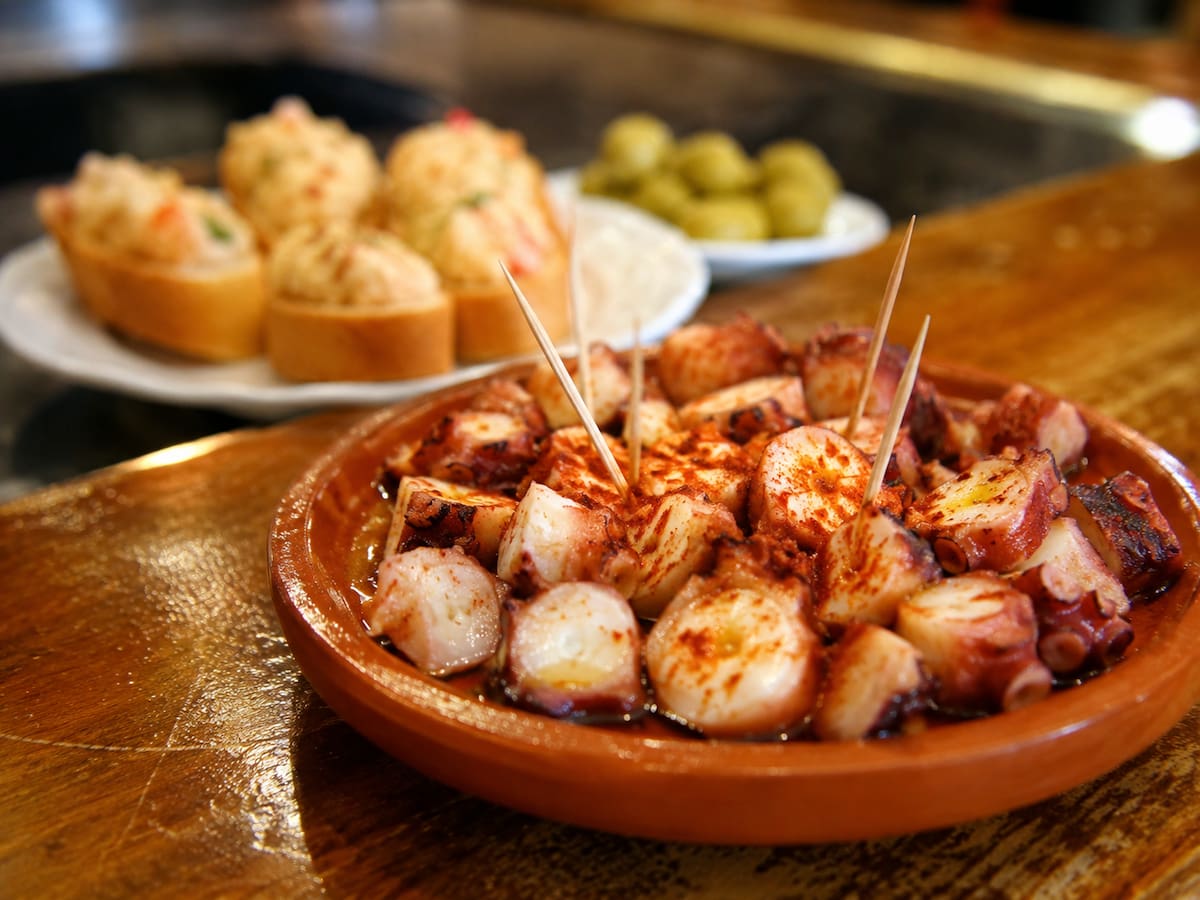 Plato de tapa española servido en un cuenco de barro con trozos de pulpo a la gallega espolvoreados con pimentón, acompañado de palillos para degustar. | Crédito: Imagen hecha con IA de ChatGPT