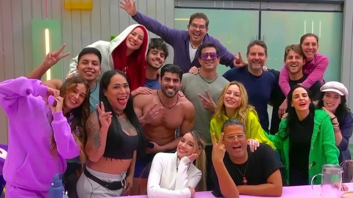 Una toma nítida y de alta resolución que captura a dieciséis participantes de un reality show posando alegres alrededor de una mesa de comedor de color rosa vibrante. La imagen muestra una amplia gama de personalidades y estilos, con gestos de victoria, poses divertidas y sonrisas genuinas. En primer plano, se aprecian detalles precisos de las pizzas artesanales y las botellas de agua sobre la mesa, mientras que el fondo revela un entorno moderno con un enrejado de madera y una gran puerta de vidrio, todo definido con gran nitidez.