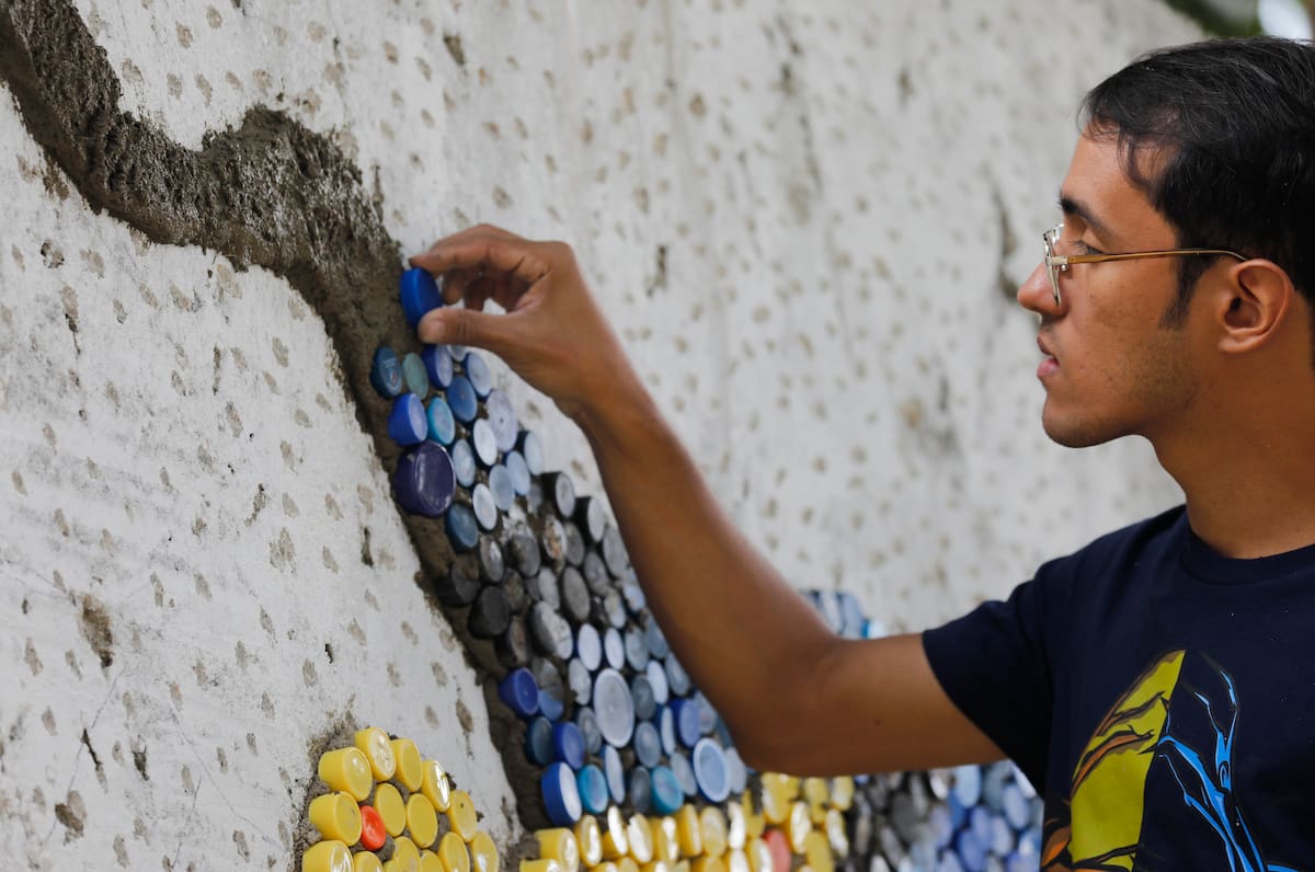 El artista venezolano Oscar Olivares coloca una tapa de botella de plástico en la pared mientras crea un mural titulado "Okomural" en El Hatillo, en las afueras de Caracas, Venezuela, el jueves 30 de enero de 2020. Olivares, de 23 años, usó miles tapas de botellas de plástico para crear este mural. (AP Foto / Ariana Cubillos)