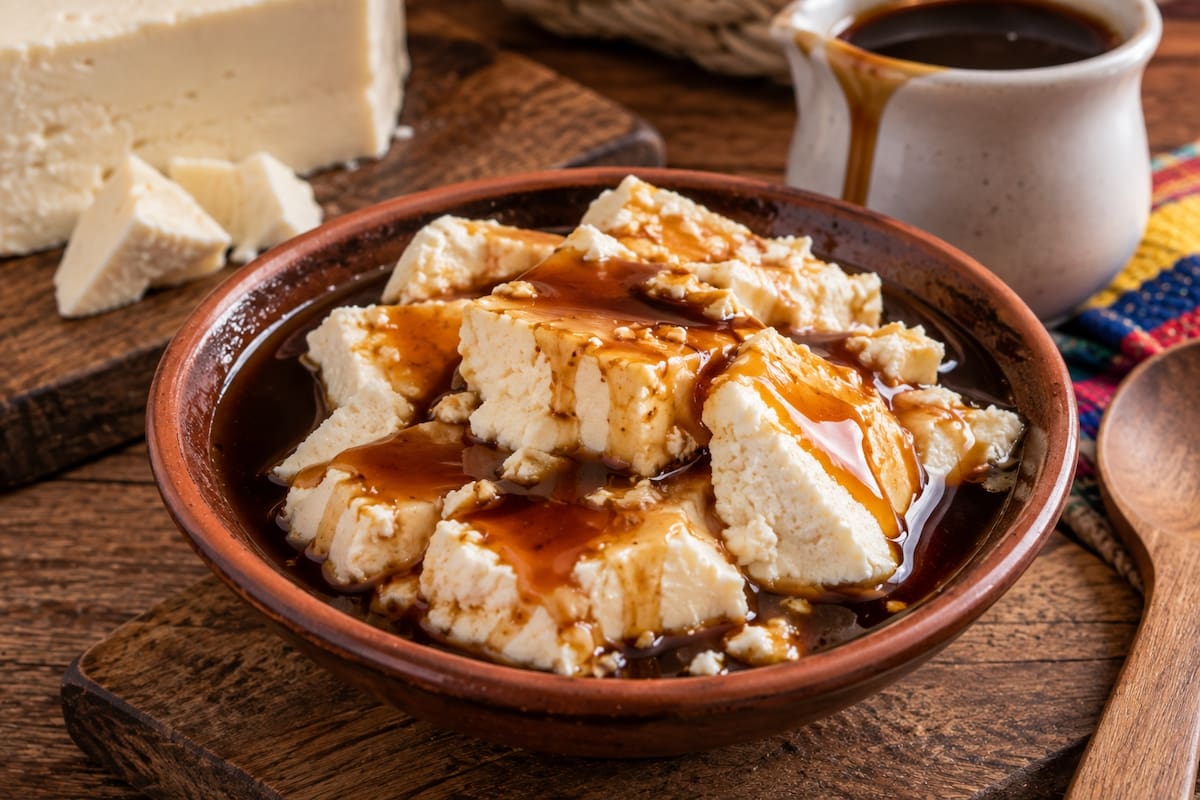 La cuajada con melao es una preparación tradicional colombiana que combina queso fresco con un jarabe dulce elaborado a partir de panela. | Crédito: Imagen hecha con IA de ChatGPT