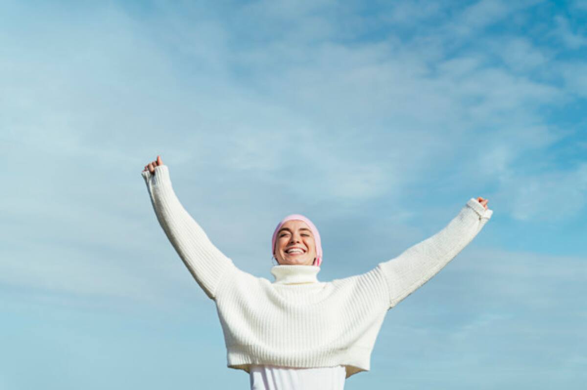 Happy young woman with cancer with arms up. She is wearing a pink scarf on her head and white jersey. She is feeling the sun on her face. She is looking to the left. Blue sky and sea on background.
