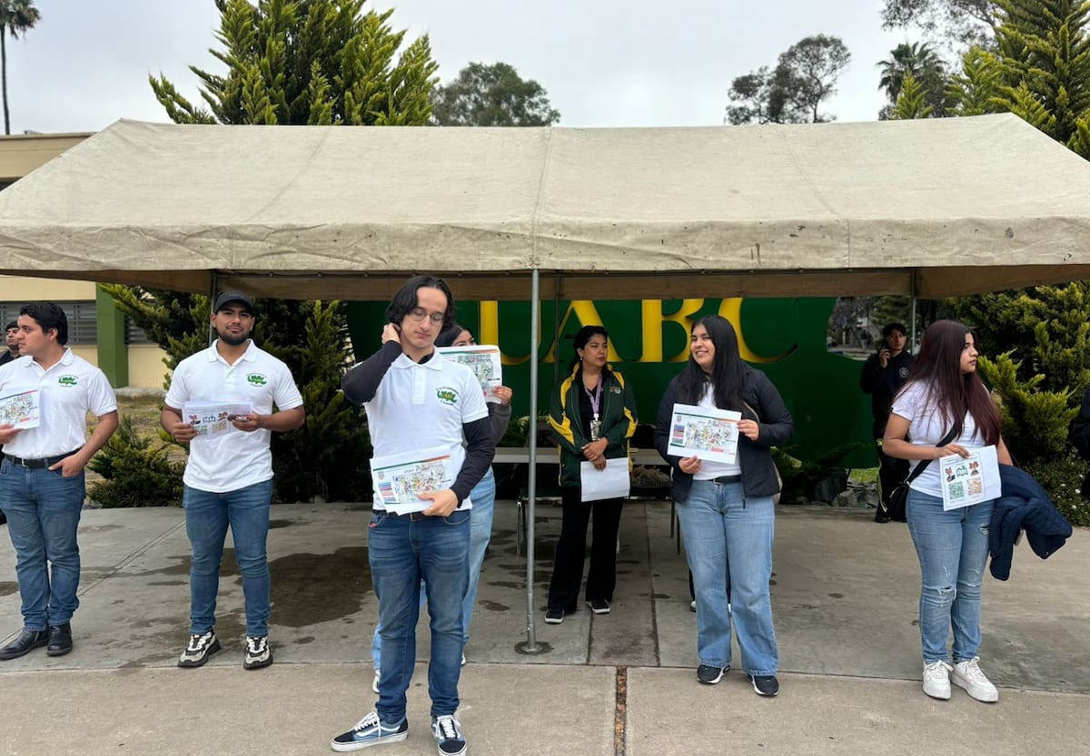 Frente al logo institucional de la UABC, estudiantes voluntarios ofrecen orientación a los aspirantes para guiarlos en la localización de sus aulas y resolver dudas generales.