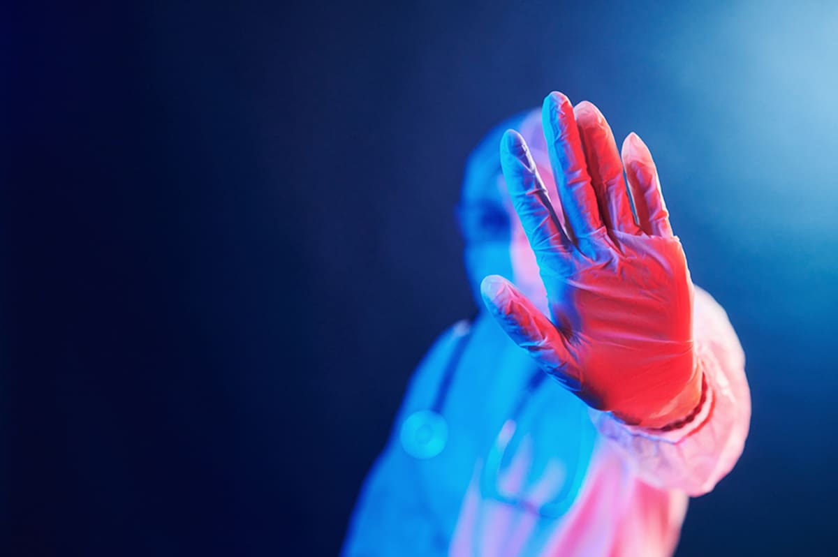 Nurse in mask and white uniform standing in neon lighted room and showing stop sign.