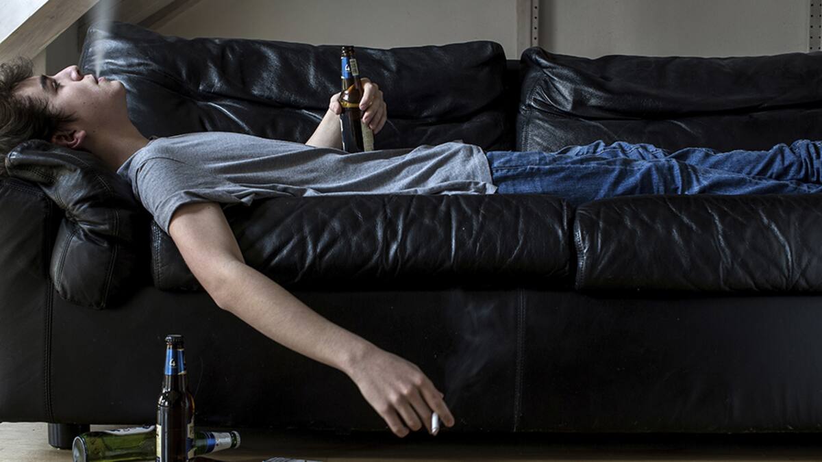 MODEL RELEASED. Young man lying on sofa holding beer and smoking cigarette.