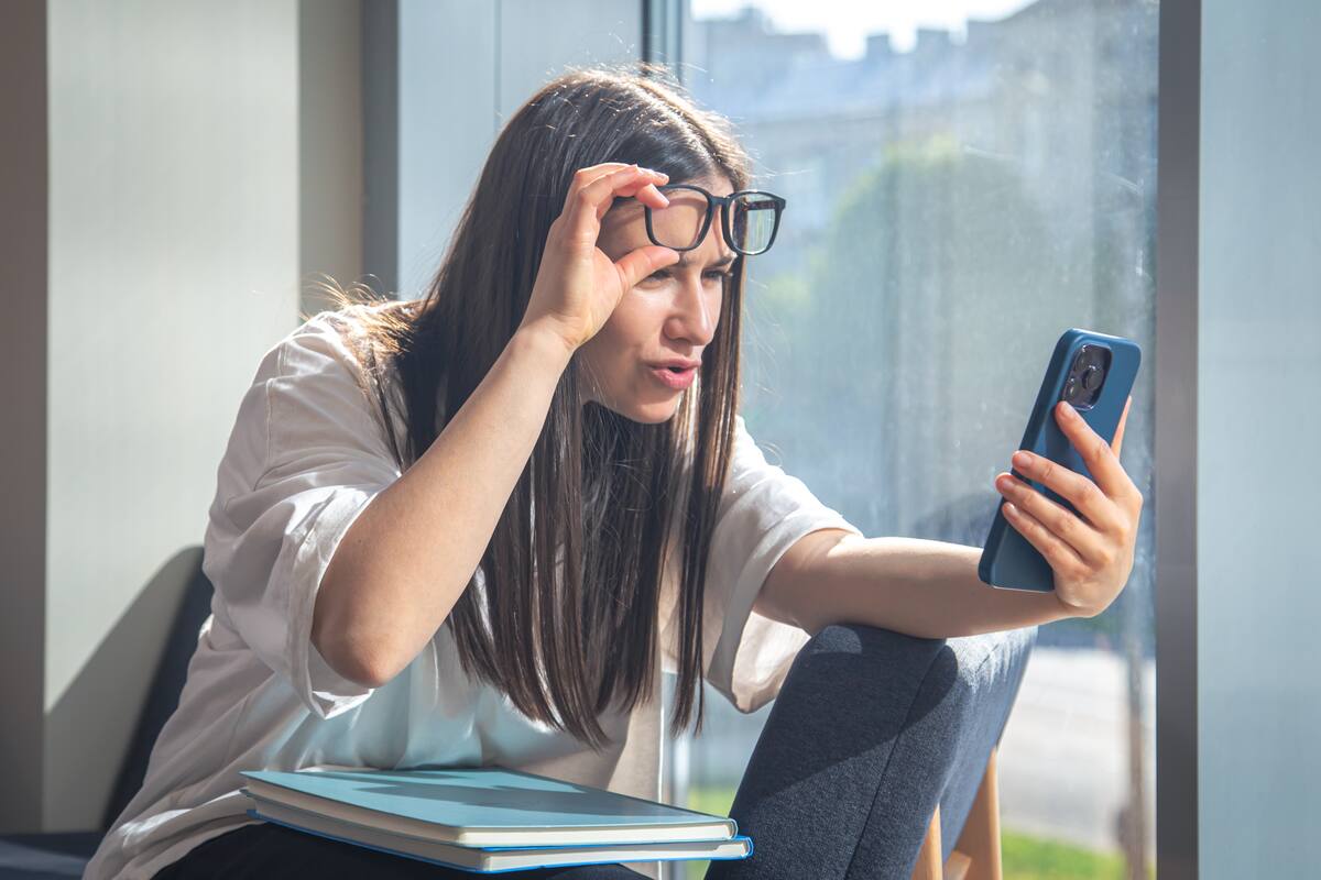 Female student looks at the screen of a smartphone in the morning by the window in surprise, the concept of study, deadline and homework.