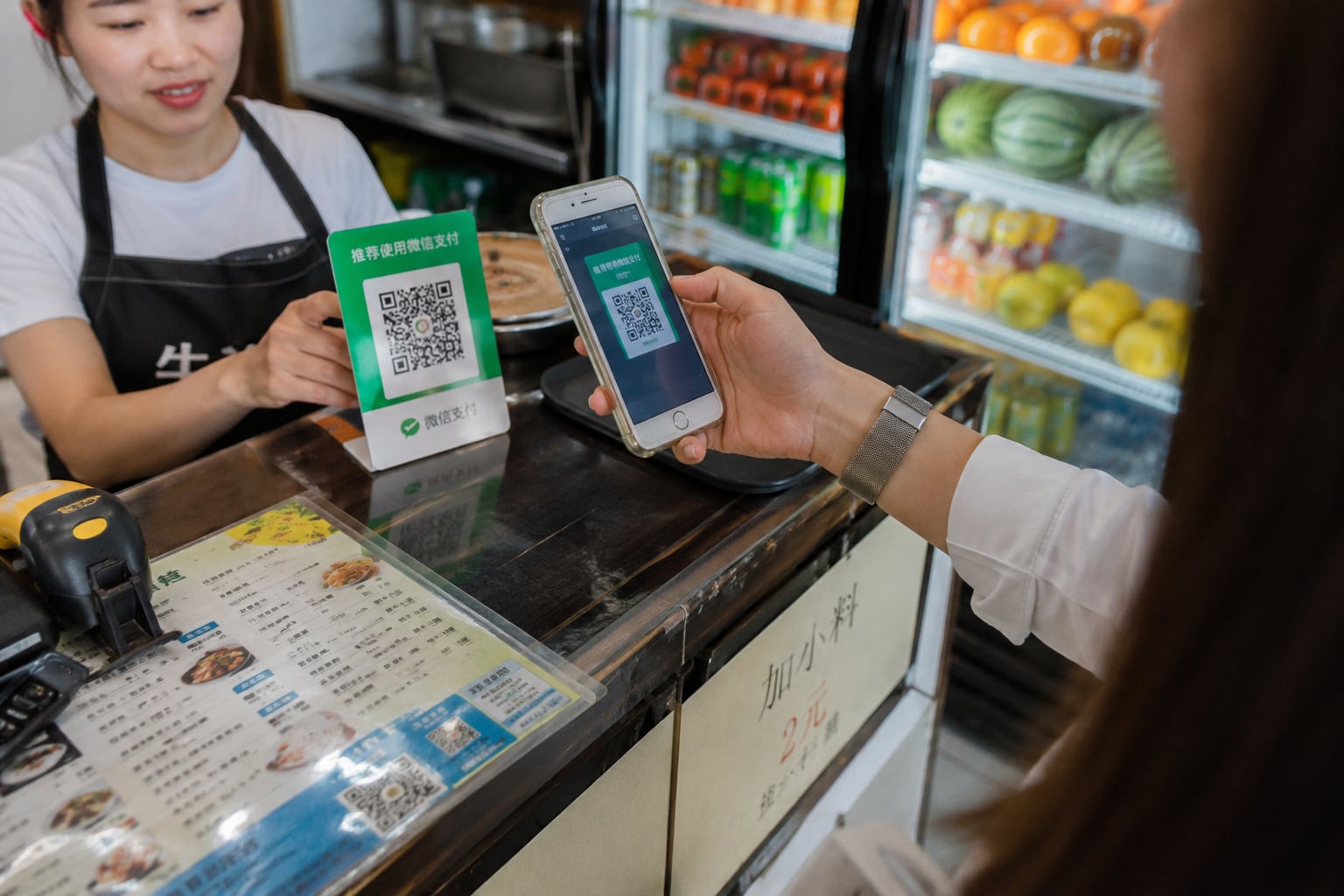 Cliente escaneando un código QR con su teléfono móvil para pagar en un pequeño puesto de comida en China, mientras la vendedora sostiene el letrero con el código; al fondo se observa una vitrina con frutas y bebidas frescas, mostrando cómo los pagos digitales dominan la vida diaria en el país. | Imagen hecha con IA de ChatGPT