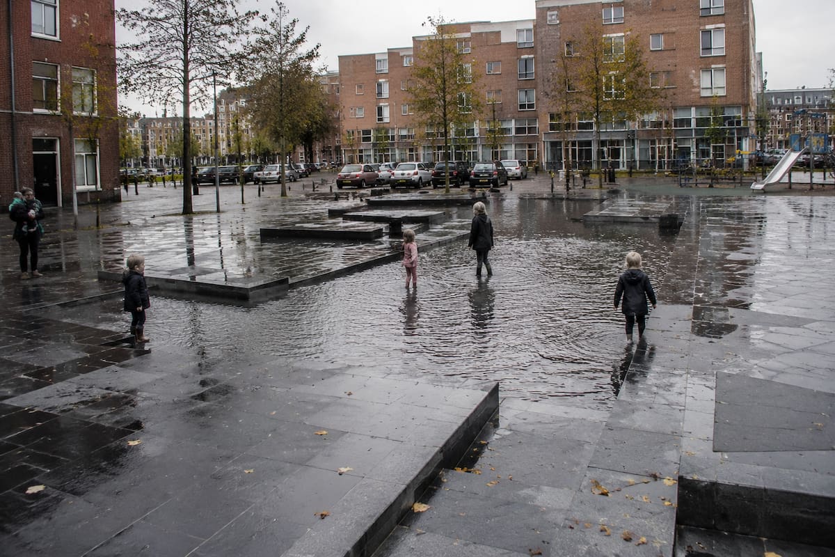 Plaza urbana en Países Bajos con “Water Squares”: charcos diseñados para acumular temporalmente agua de lluvia, donde niños juegan mientras la infraestructura combina funcionalidad y espacio público seguro. | Imagen hecha con IA de ChatGPT